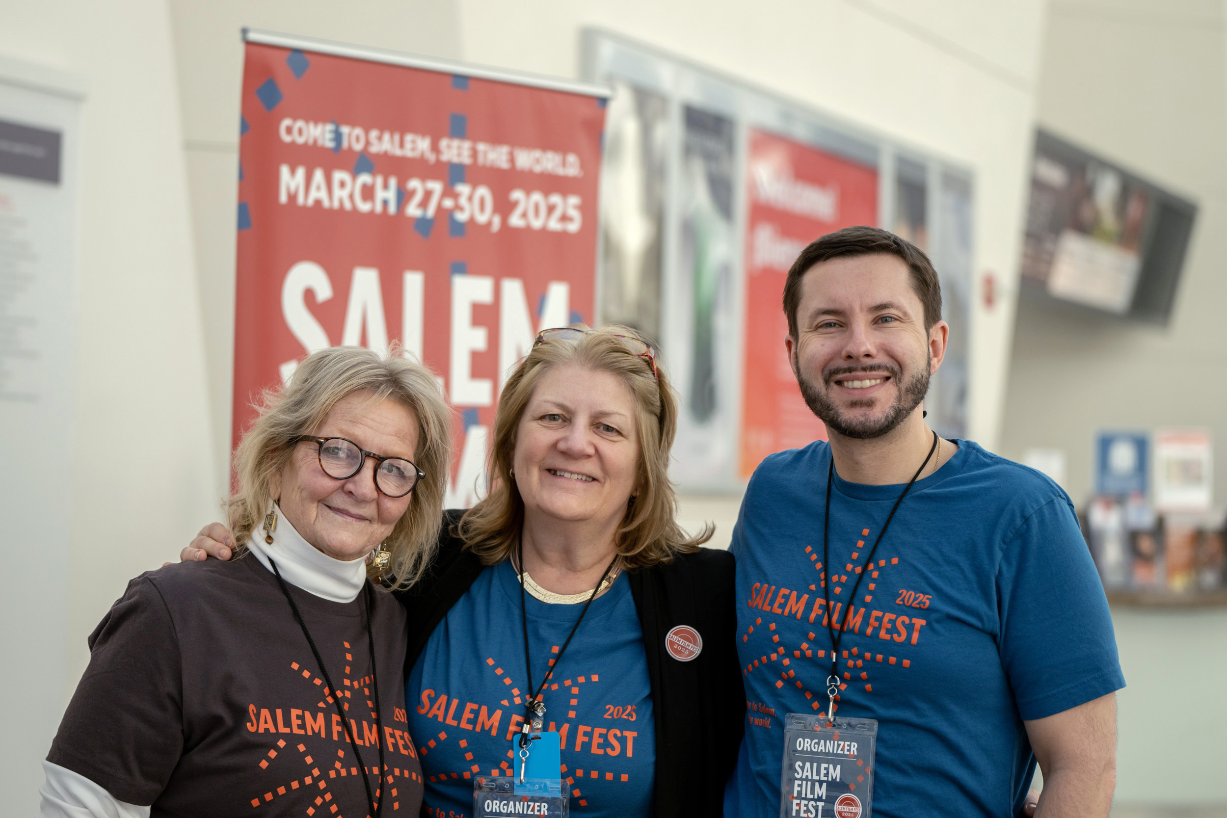 Three smiling Salem Film Fest organizers unite indoors, badges and shirts on, celebrating creativity under a bold local event banner.