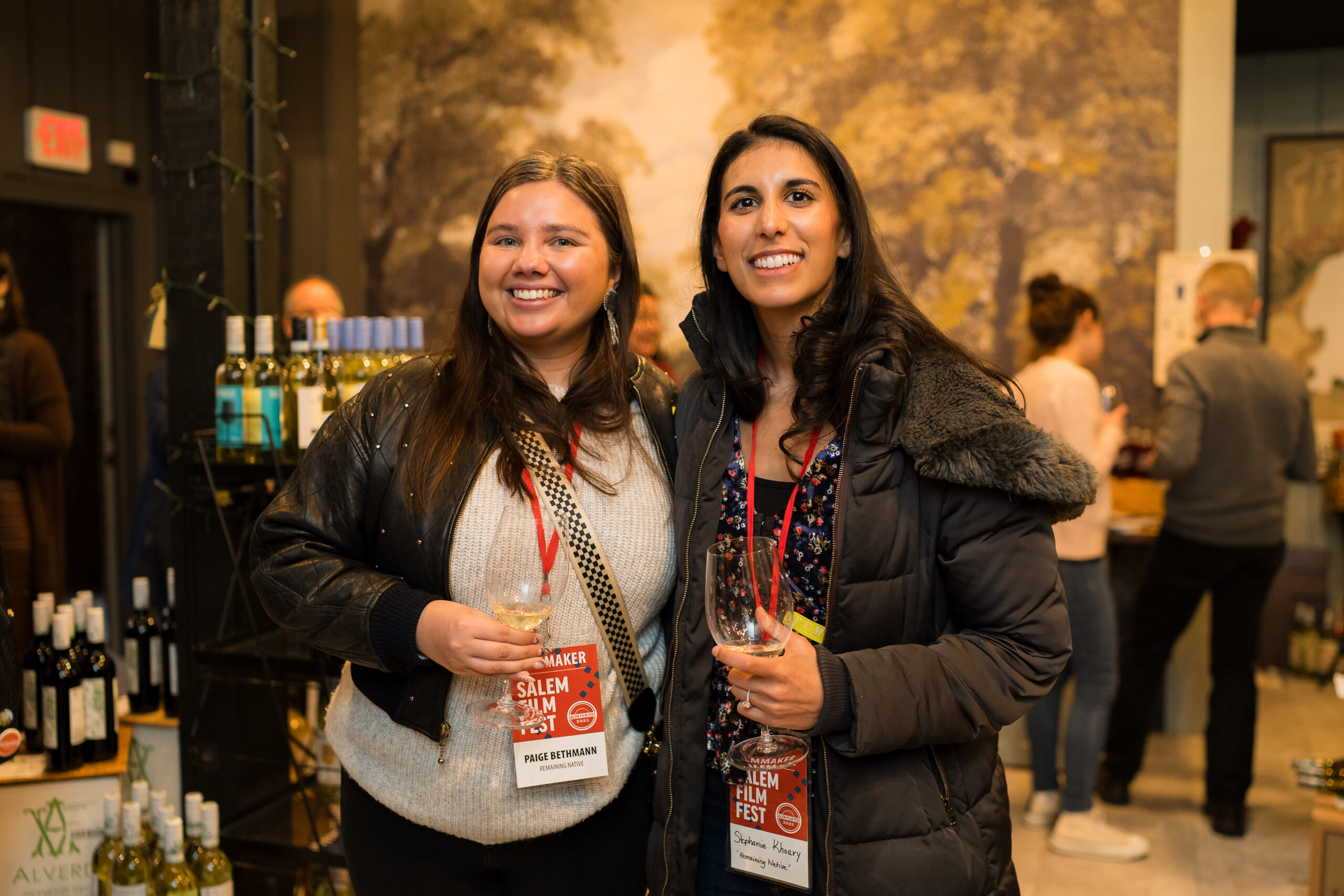 Two women in Salem Film Fest badges toast with wine, surrounded by buzzing locals and shelves of Essex County’s creative energy.