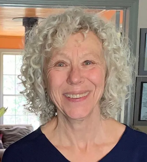 An older woman with short, curly gray hair is smiling while standing indoors. She is wearing a dark blue top, and there is a window and framed art in the background, highlighting Essex County Artists recognized by 2026 Massachusetts Cultural Council Grants.