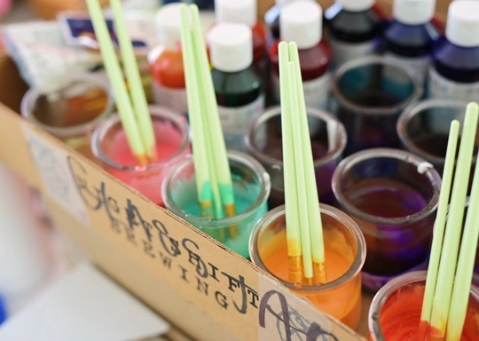 Clear cups filled with different colored liquids, each holding two paintbrushes, are arranged in a cardboard container—perfect for a K-2nd Art Design Club. Bottles of vibrant colorants or dyes are visible in the background.