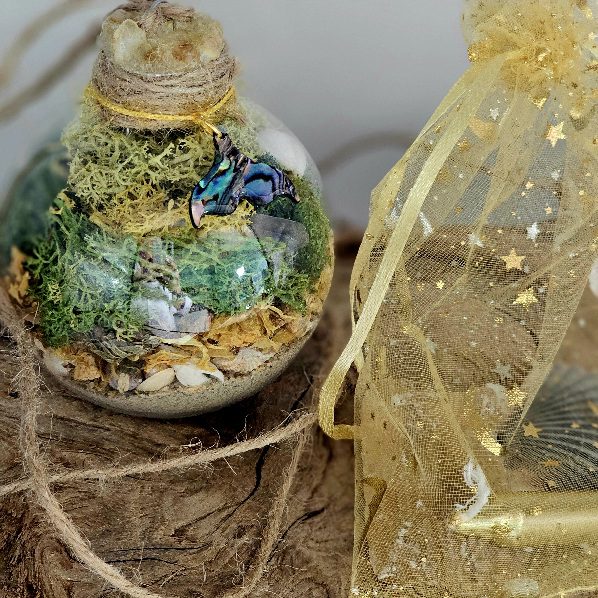 A glass terrarium ornament filled with layers of sand, small stones, green moss, and a decorative butterfly sits on a textured surface next to a translucent yellow drawstring bag with star patterns.