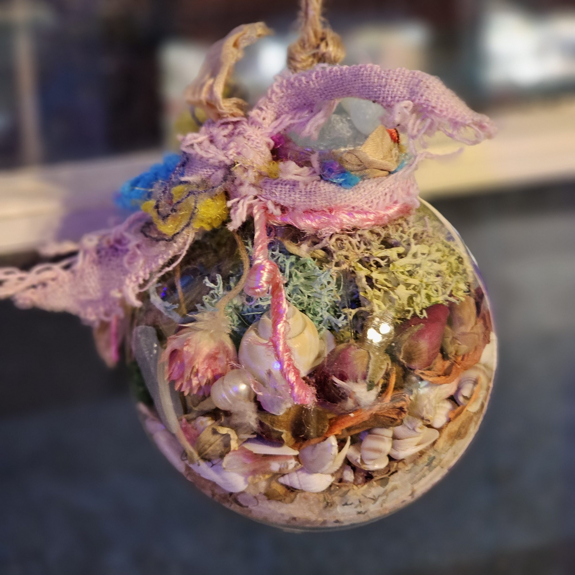 A clear ornament filled with small seashells, dried flowers, moss, and other natural materials, topped with a pink and purple frayed cloth and twine, hanging against a blurred background.