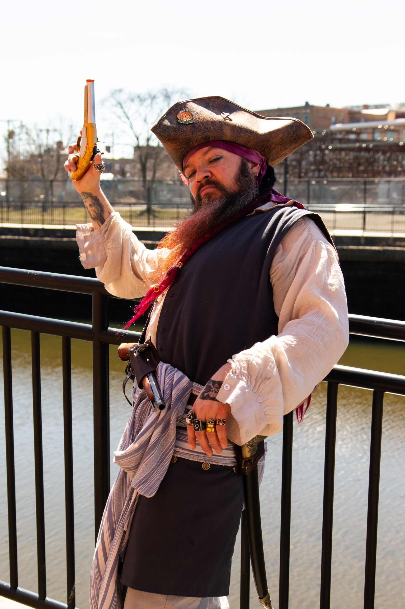 A person dressed as a pirate poses outdoors by a railing, holding a yellow toy pistol. Channeling Salem’s maritime history, they wear a tricorn hat, white shirt, dark vest, striped sash, and sport accessories and visible tattoos.