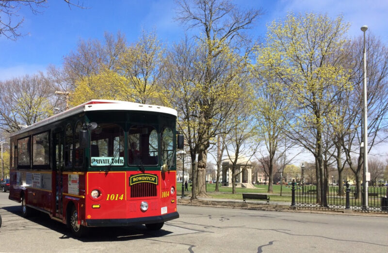 A red and green trolley labeled "Bowditch" with the number 1014, part of the Salem Trolley, drives on a street next to a park where trees are just starting to bud. Benches and a fence appear in the background under a blue sky.