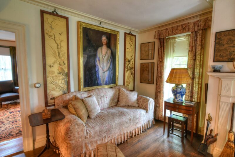 A floral upholstered sofa sits beneath a large framed portrait in a traditional living room with wooden floors, patterned curtains, a side table with a blue and white lamp, and decorative wall panels.