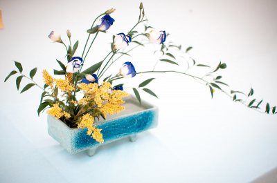 A rectangular ceramic vase with a blue glaze holds an arrangement of yellow and blue flowers, along with green leafy stems, displayed on a white surface.