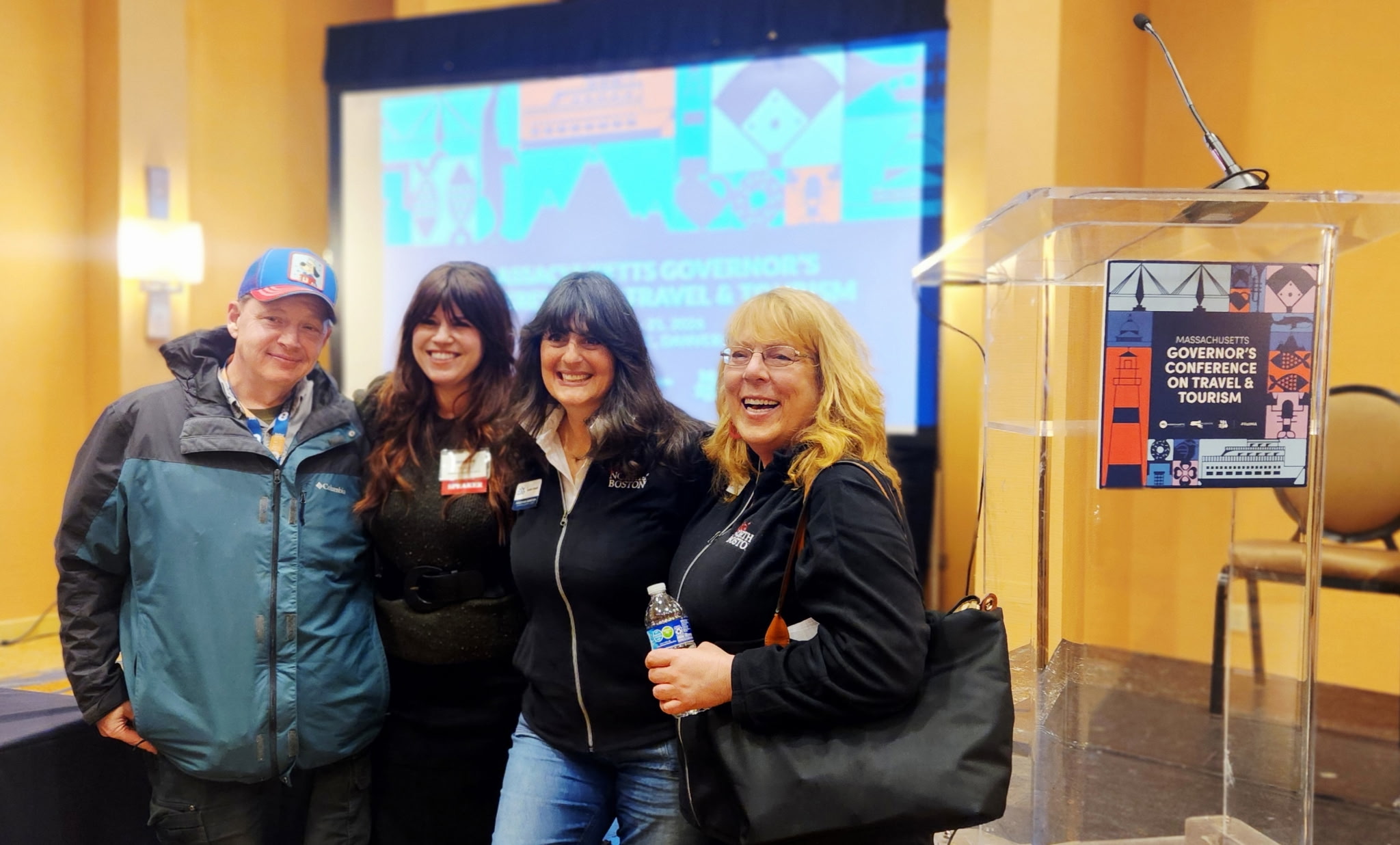 Four people stand together smiling in front of a podium and a screen displaying a presentation on arts and culture at a conference. They are wearing casual and business-casual clothing with conference badges.