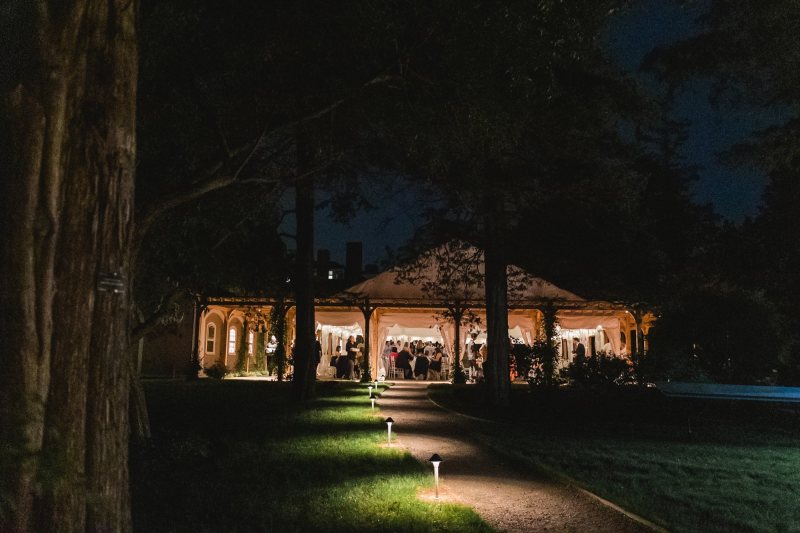A view of a well-lit pavilion at night, surrounded by trees and a grassy lawn, with several people gathered inside for the Preview Party. A pathway with small lights leads from the foreground to the building.