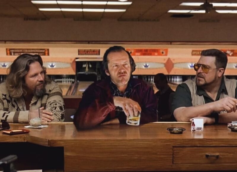 Three men sit at a bowling alley bar; the man on the left wears a beige sweater, the man in the middle holds a drink, and the man on the right sports sunglasses and a vest, exchanging trivia as bowling lanes stretch out behind them.
