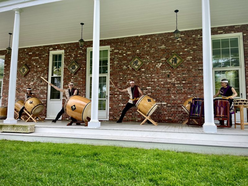 Four people play large traditional drums on a covered porch at Long Hill, in front of a brick building with tall windows. Dressed in matching black outfits and headbands, they fill the air with rhythms inspired by Japan. The event offers general admission seating.
