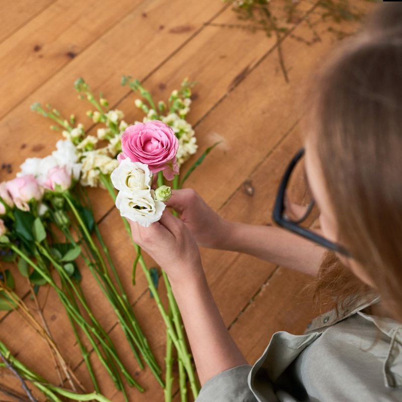 A person with glasses arranges pink and white flowers on a wooden surface during a Floral Workshop, holding a small bouquet while several loose stems lie nearby.