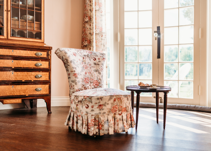 A floral upholstered chair sits next to a small round table in front of glass-paned double doors. Reminiscent of New England servant life, a wooden cabinet with drawers stands on the left as sunlight streams in through the doors.