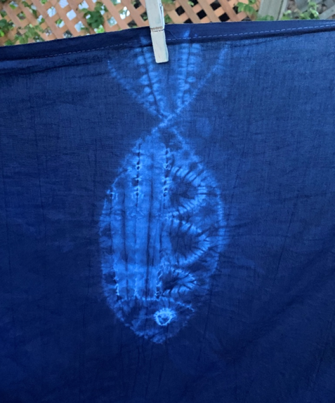 A dark blue fabric with a light blue tie-dye pattern resembling a stylized fish shape, created at an Indigo Shibori Workshop in Japan, hangs from a clothesline with a wooden clothespin. A wooden lattice and greenery are visible in the background.