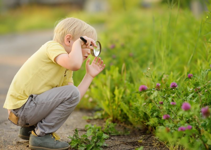 A young child with blond hair squats outdoors, holding a magnifying glass and closely examining green plants and purple flowers during a spring outdoor adventure in a grassy area.