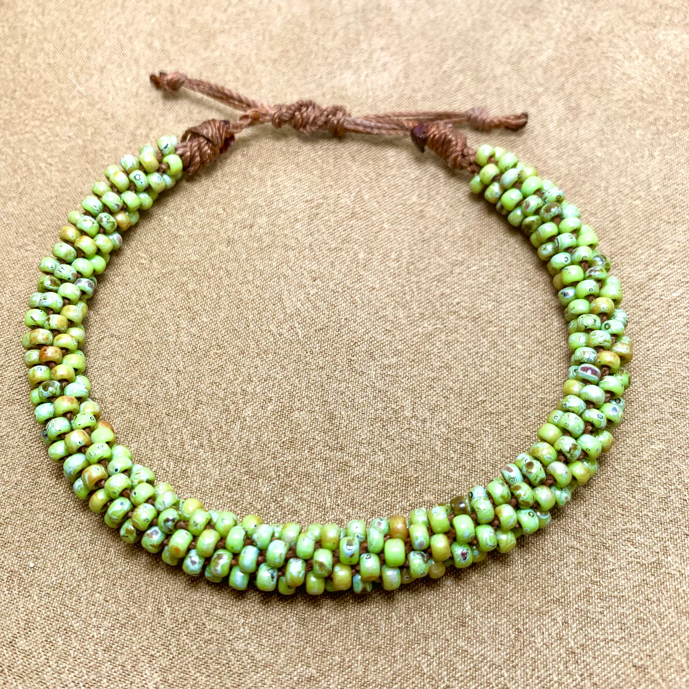 A beaded choker necklace with green and brown speckled beads strung closely together on a brown cord, inspired by Kumihimo techniques, displayed on a textured tan surface.