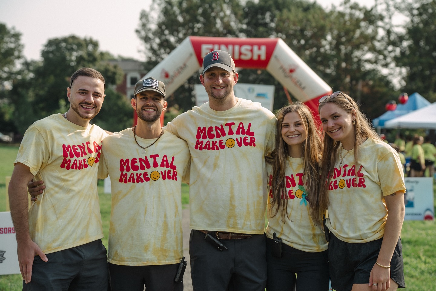 Five people stand together outside, smiling and wearing matching yellow "Mental Makeover" t-shirts at the 5th Annual Walk. They are in front of a large inflatable arch that says "FINISH," with trees and tents in the background.