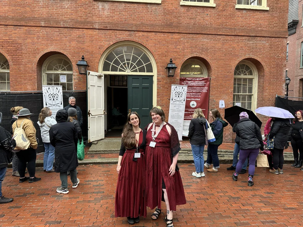 Two women in matching maroon dresses smile in front of a historic brick building with arched windows at the 2026 Fiber Witch Festival in Salem. Several people with umbrellas and coats stand in line outside on a wet day.