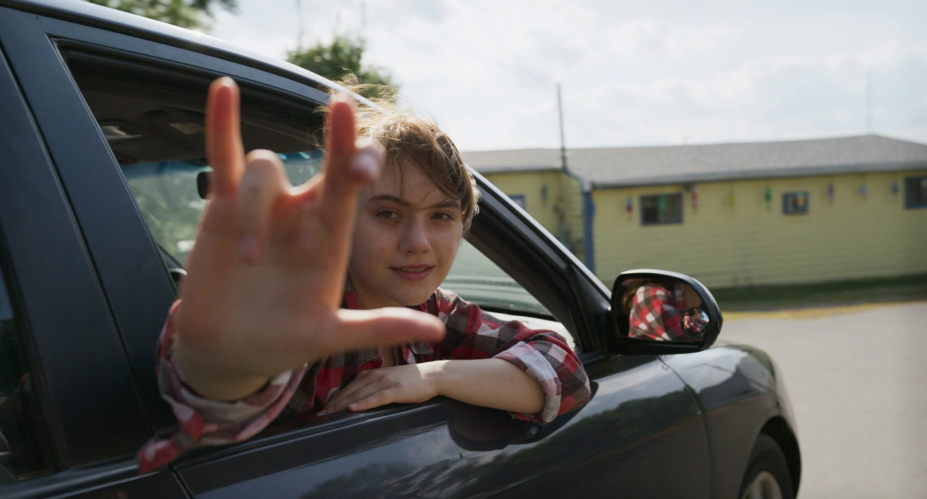 A person in a plaid shirt sits in a car with the window down, extending their arm outside and making the “I love you” hand sign in American Sign Language. A yellow building is visible in the background.
