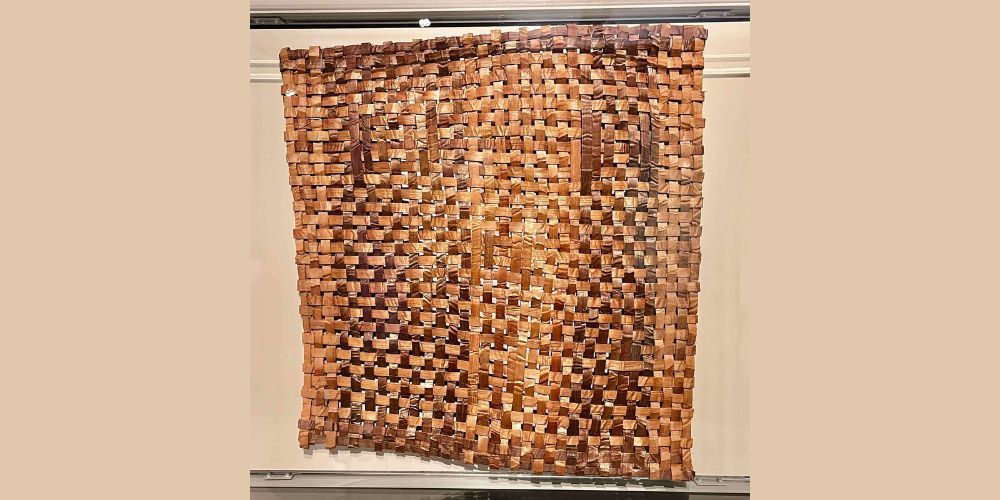 A rectangular woven mat made of brown and tan strips of material hangs vertically against a light-colored wall. The strips are interlaced, creating a varied checkerboard pattern.