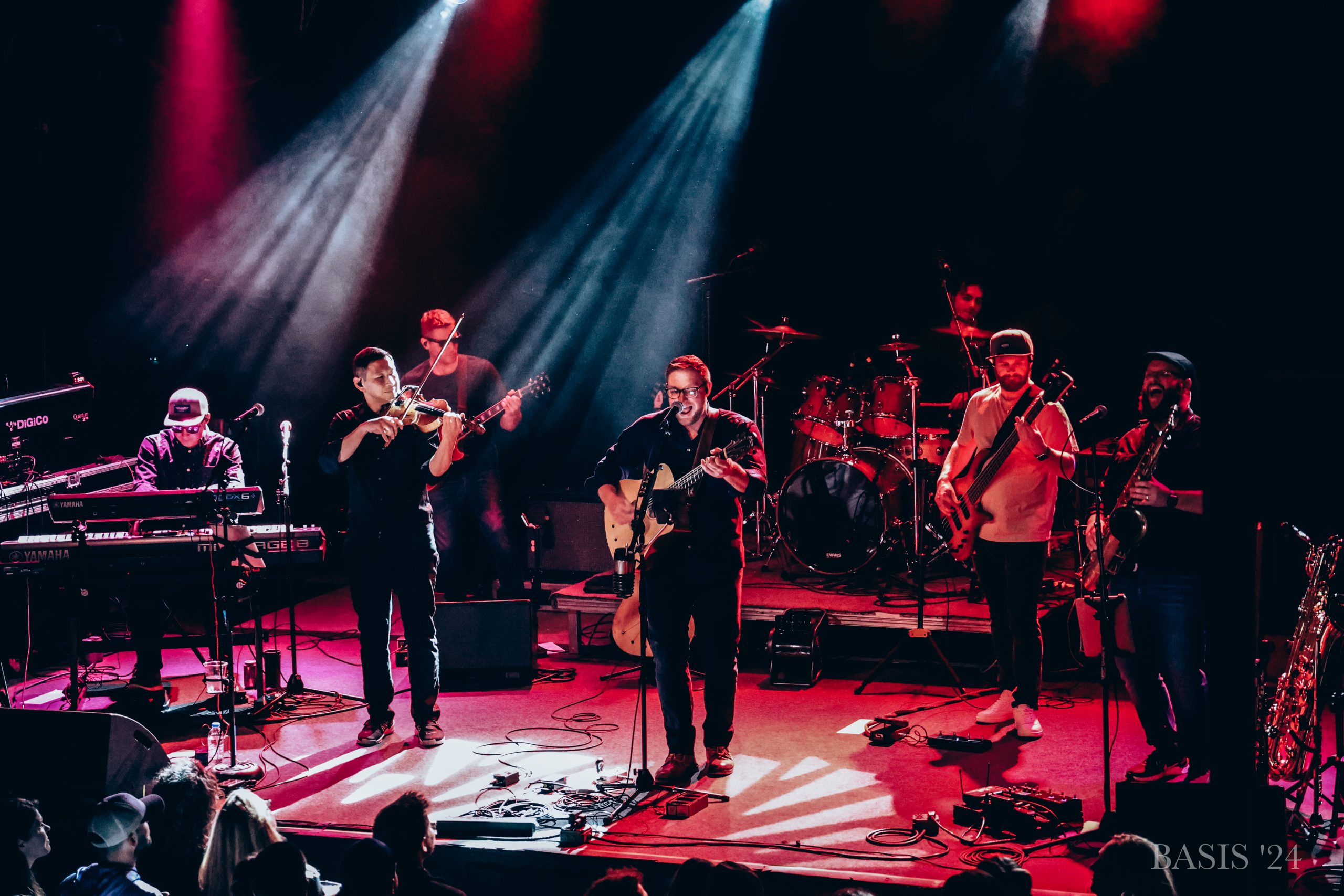 A band of seven musicians performs on stage under colorful lights, playing various instruments including keyboard, violin, guitar, drums, bass, and brass, with an audience visible in the foreground.