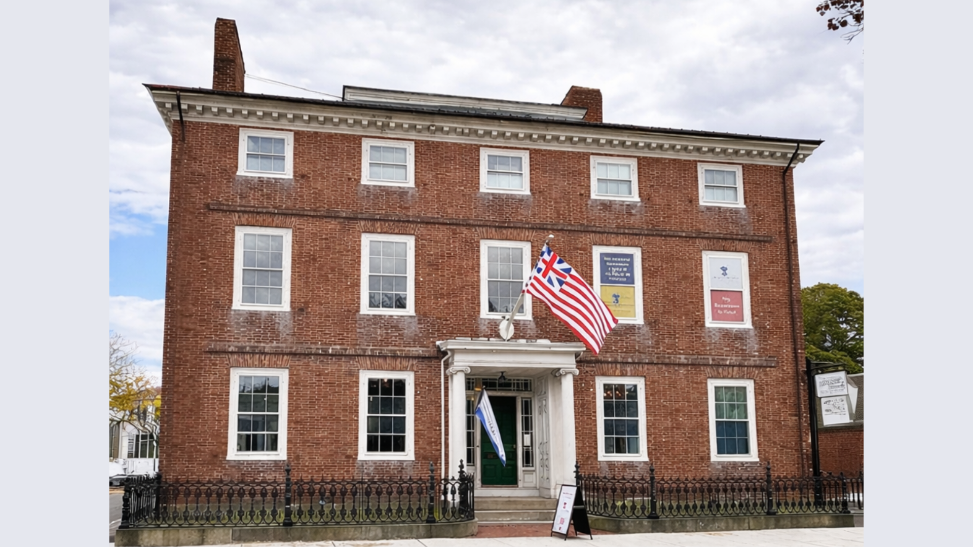 A three-story red brick building with white-trimmed windows, two flags above the entrance, and several plaques on the facade stands under a cloudy sky. A black iron fence lines the front, marking this site as part of Historic Beverly.