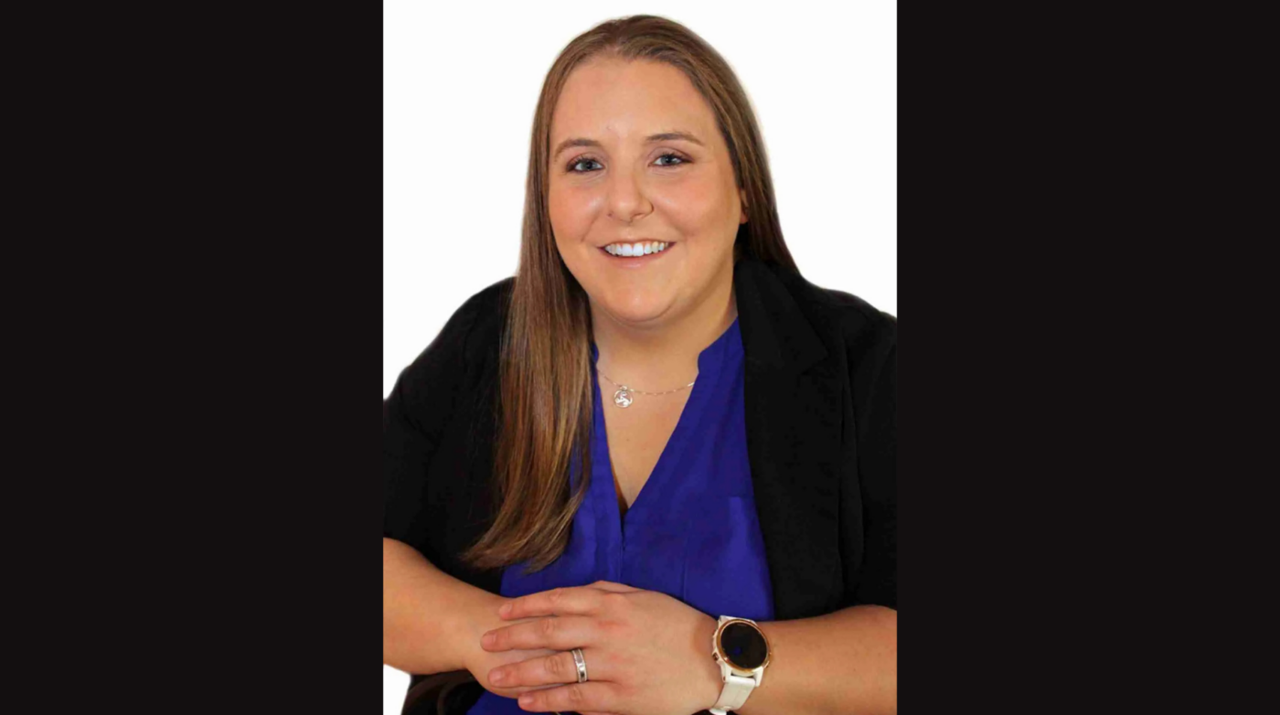 Katie Kimball Dyer, a woman with straight, light brown hair, smiles at the camera. She is wearing a blue blouse, black blazer, white watch, and a necklace, with her hands folded in front of her against a white background.