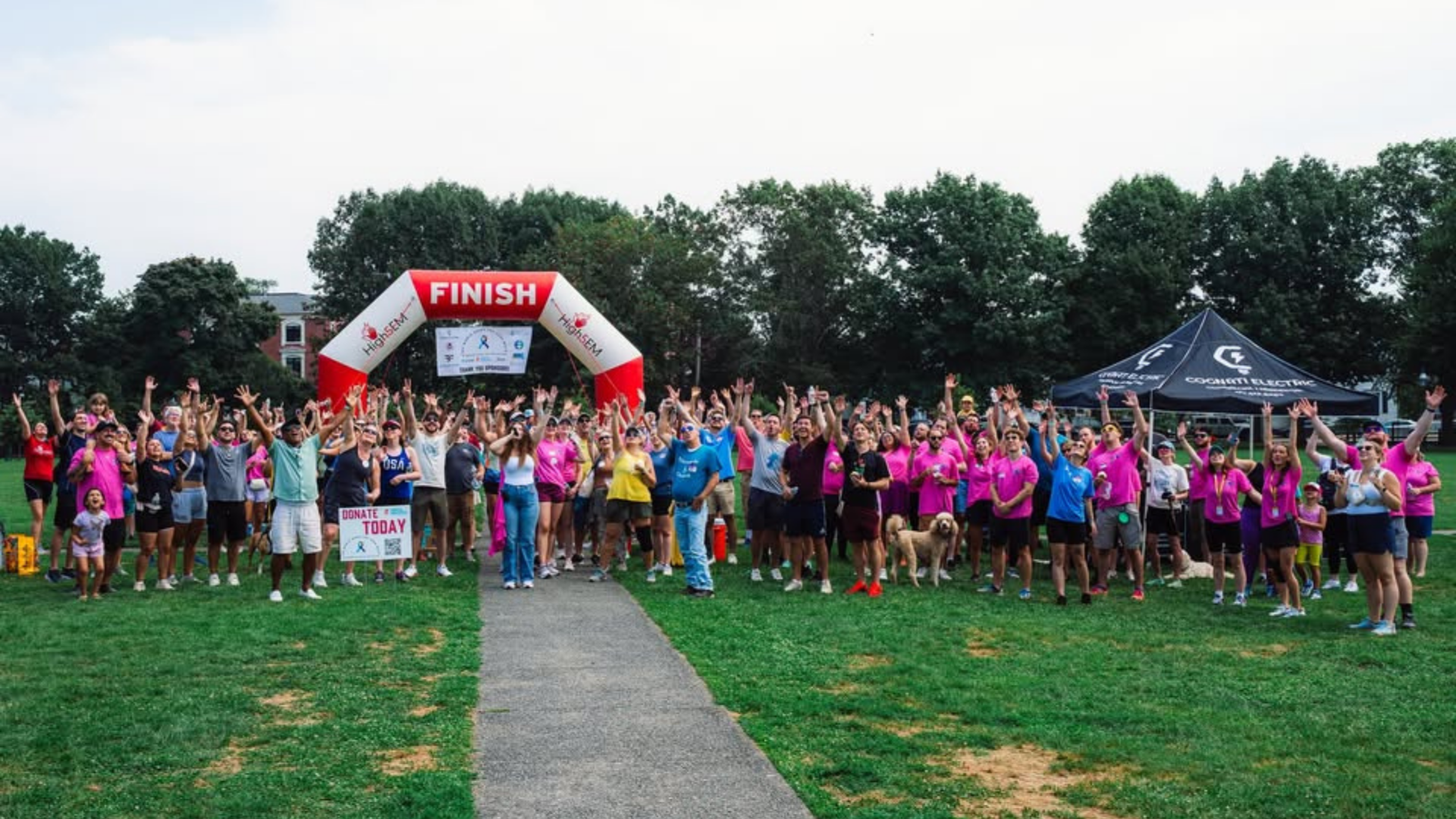 A large group of people, including members of the North Shore AAPI Coalition, pose with raised arms beneath a red and white finish line arch on a grassy field, with trees and tents in the background. Many participants wear pink shirts.