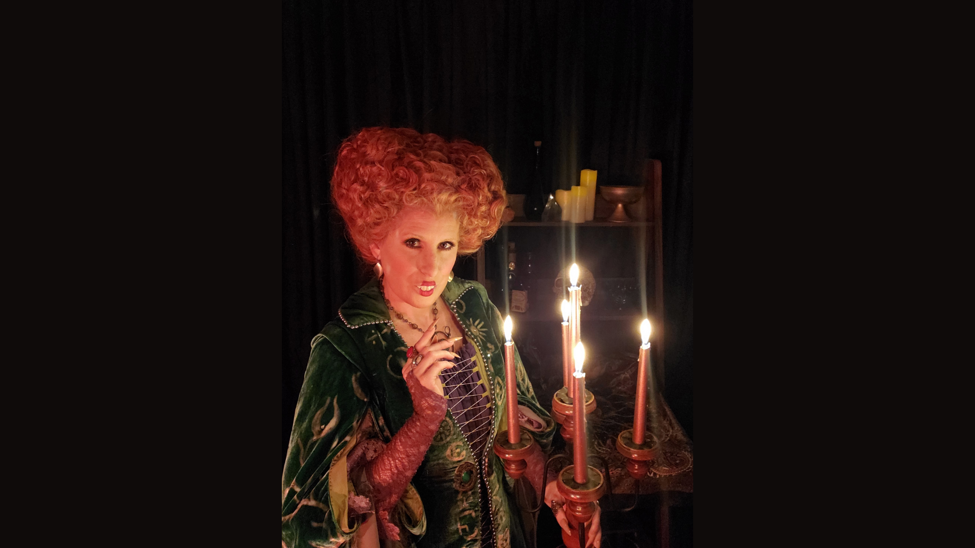 A person in a green velvet costume with elaborate red hair stands indoors beside a candelabra with four lit candles. Meet the Member: Wicked Makeup brings a dramatic flair to the dark room, where yellow candles and shelves are visible in the background.