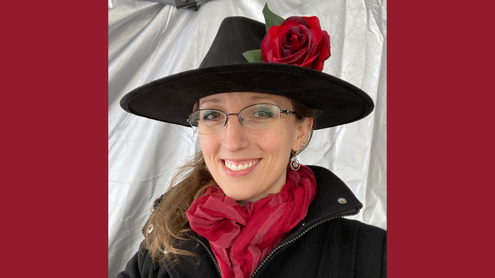 Meet the Member: Mental Makeover—a person wearing glasses, a black wide-brimmed hat with a red rose, a black coat, and a red scarf, smiling in front of a gray background.