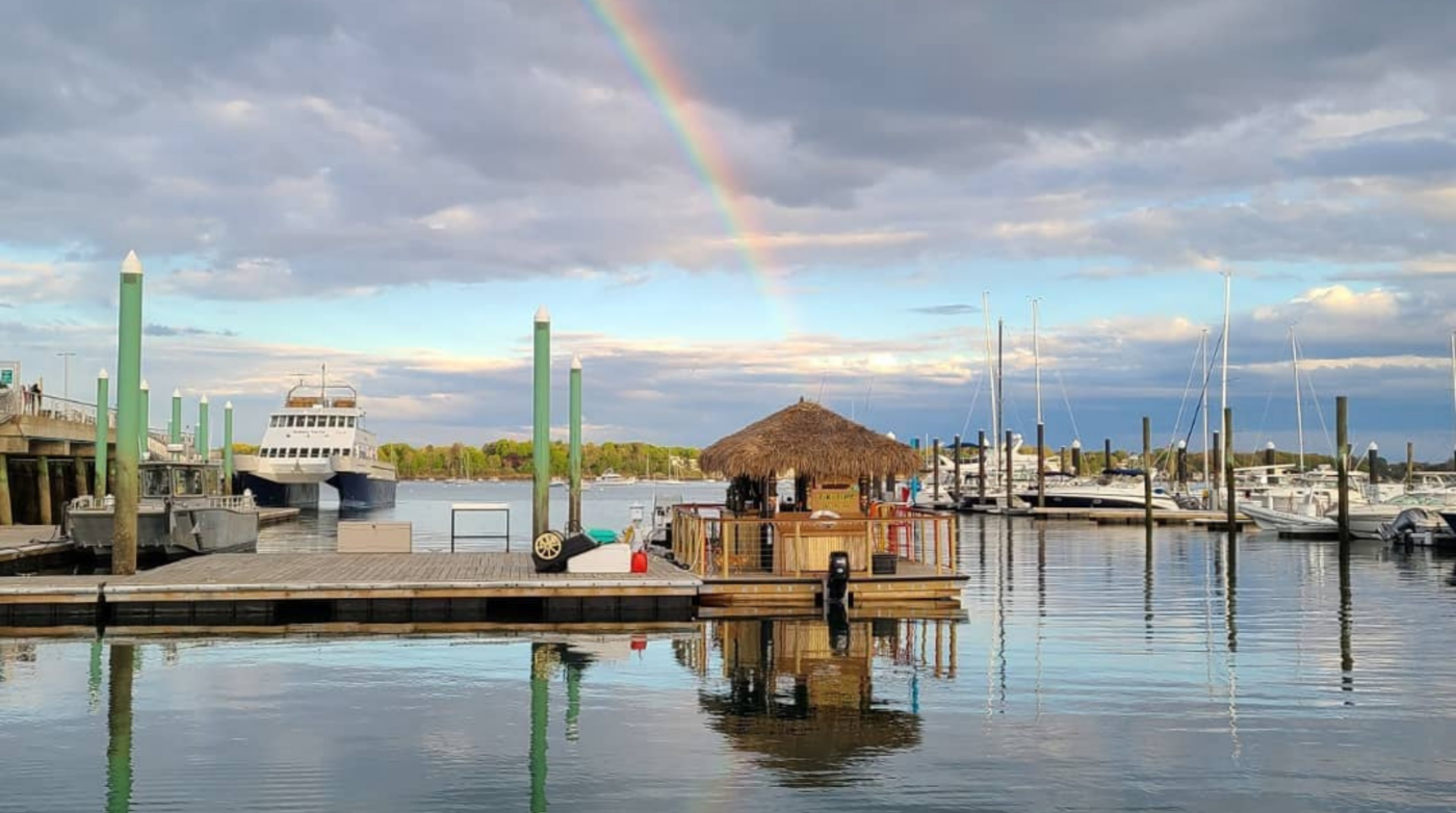 A marina with several boats docked, including Tiki Hut Boats on a floating platform. Calm water reflects a rainbow arched in the cloudy sky, with some trees visible in the background.