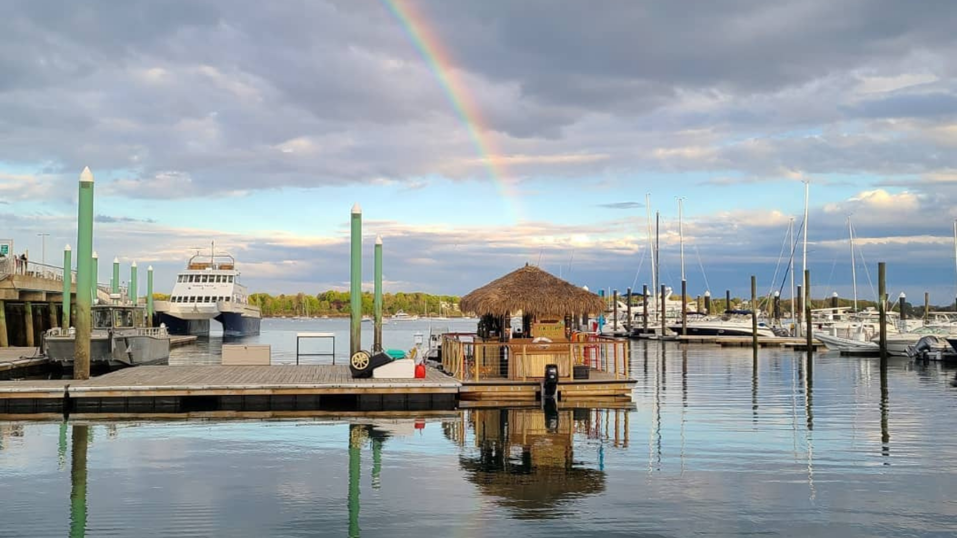 A marina with several boats docked, including Tiki Hut Boats on a floating platform. Calm water reflects a rainbow arched in the cloudy sky, with some trees visible in the background.