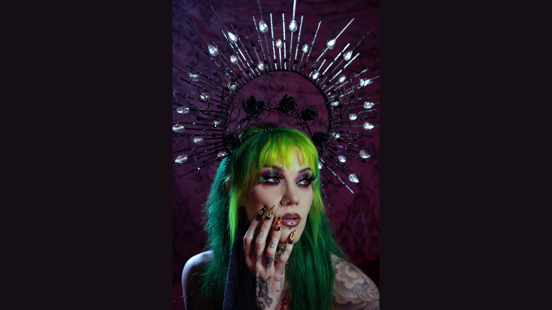 Meet the Member: Sanazar Rose—a person with long green hair and dramatic makeup wears a black spiked crown adorned with crystal-like decorations. Their tattooed hand with ornate nails rests on their face against a dark, textured background.