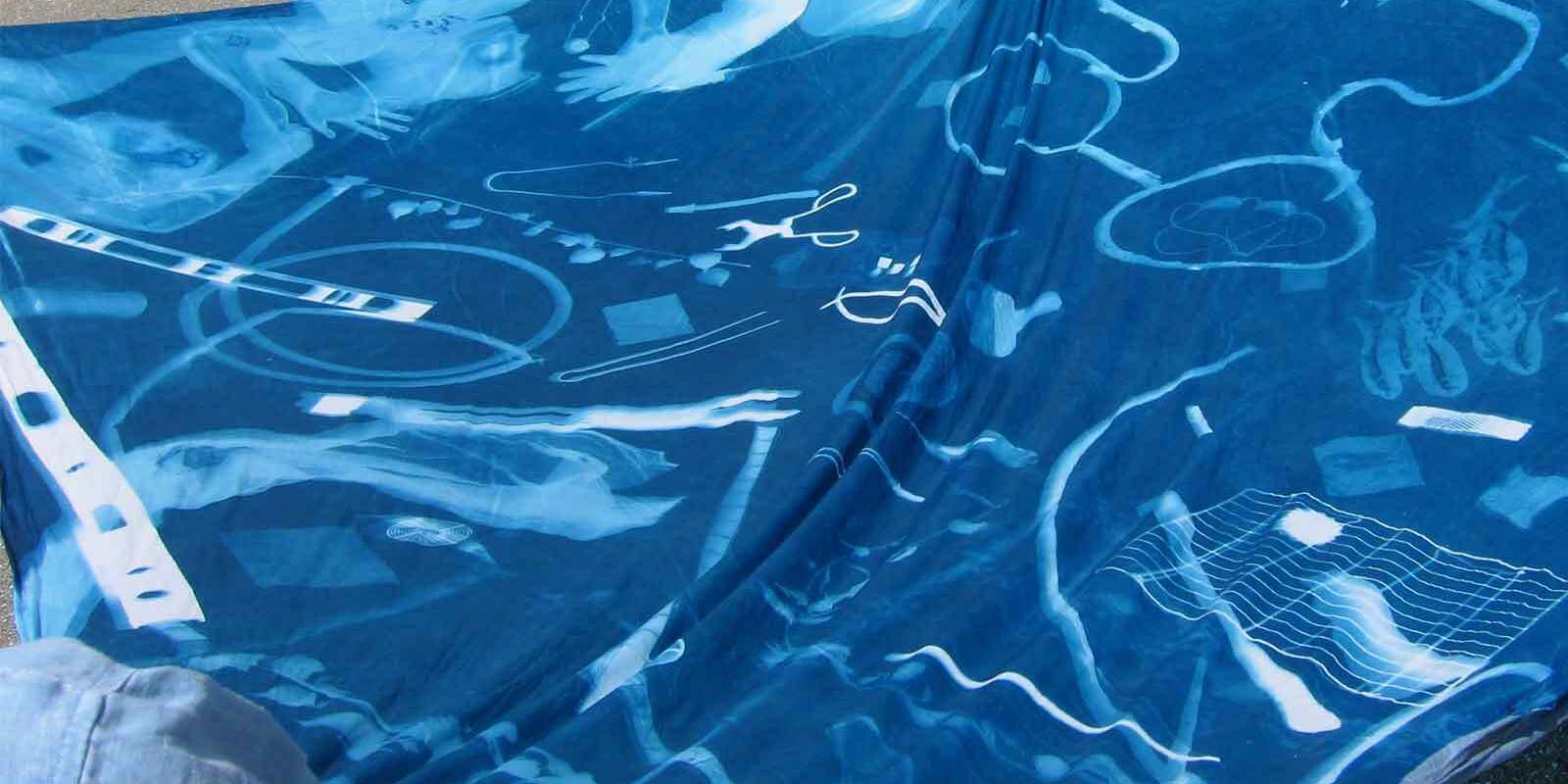 A large blue fabric displays white silhouettes of various objects, including tools, rings, wires, and geometric shapes, resembling a cyanotype print laid out on a flat surface.