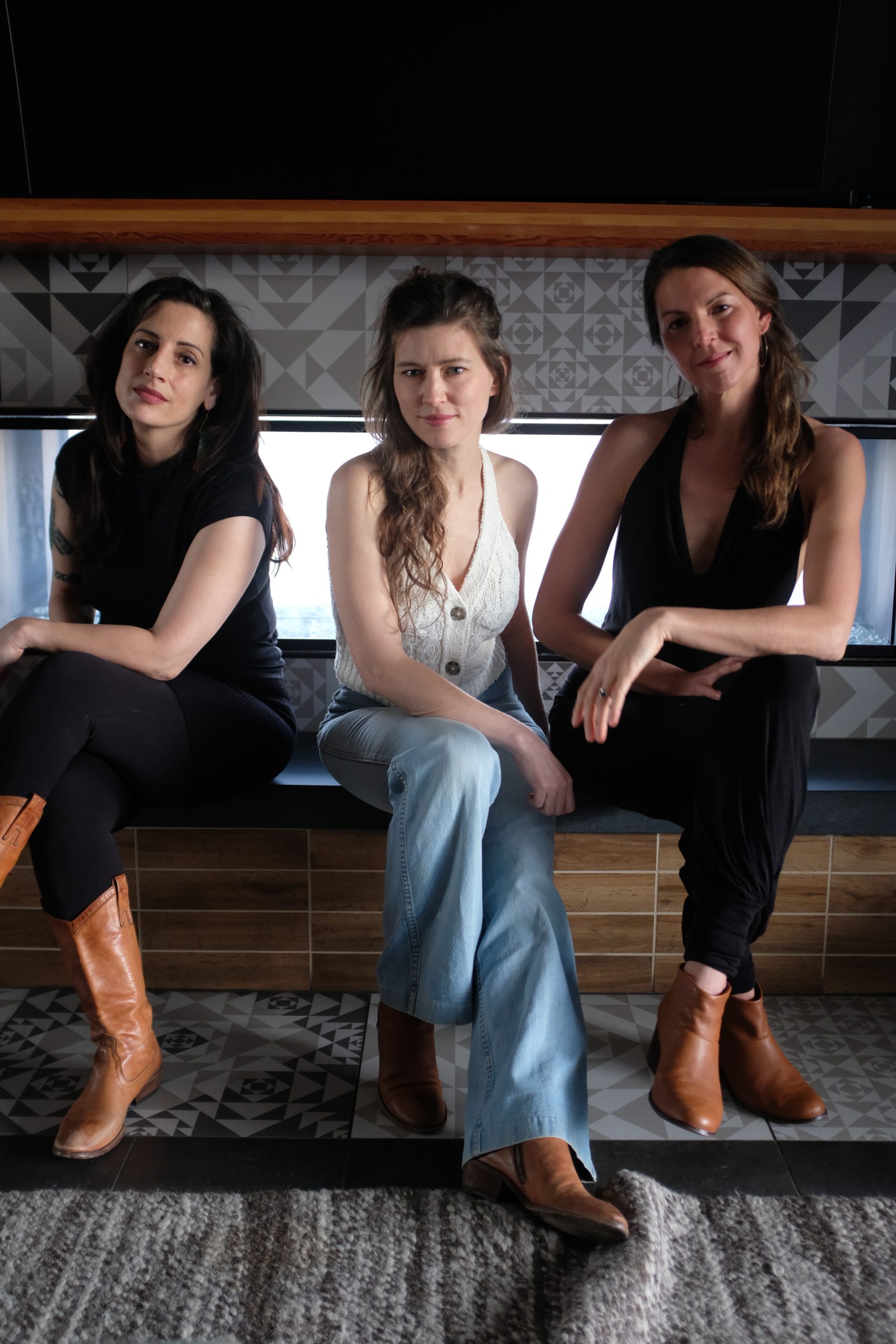 Three women sit side by side on a bench with patterned tiles behind them. They wear casual outfits and brown ankle boots, looking toward the camera with neutral expressions.