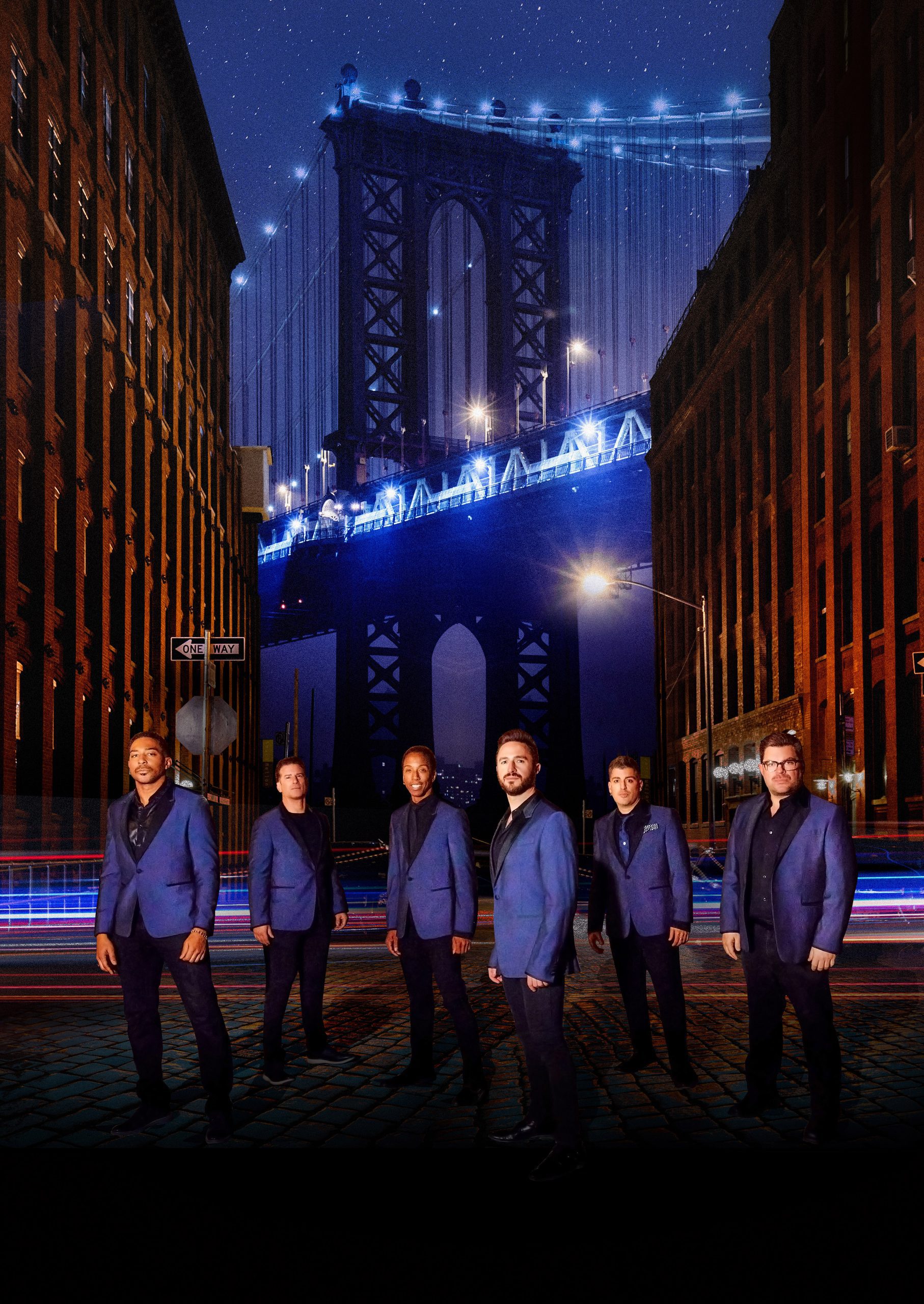 Six men wearing blue jackets stand on a cobblestone street at night, with the Manhattan Bridge illuminated in the background and tall buildings on either side. Light trails from vehicles are visible.