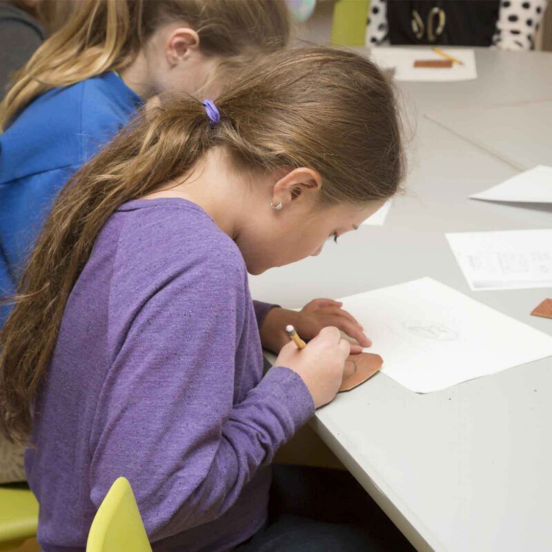 A young girl with long brown hair tied back, wearing a purple shirt, sits at a table and draws on a piece of paper with a Tiny Stick pencil. Other children are seated nearby, working on their projects and decorating Window Picture Frames.