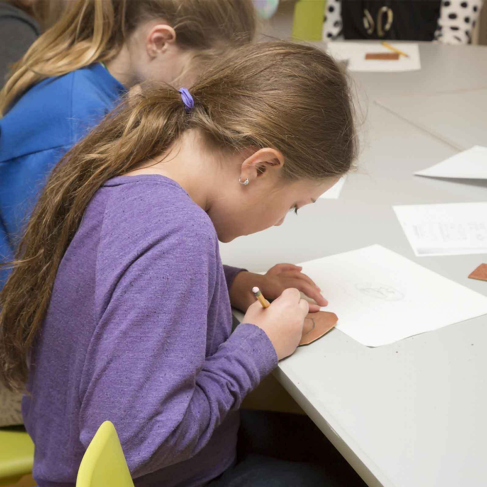A young girl with long brown hair tied back, wearing a purple shirt, sits at a table and draws on a piece of paper with a Tiny Stick pencil. Other children are seated nearby, working on their projects and decorating Window Picture Frames.