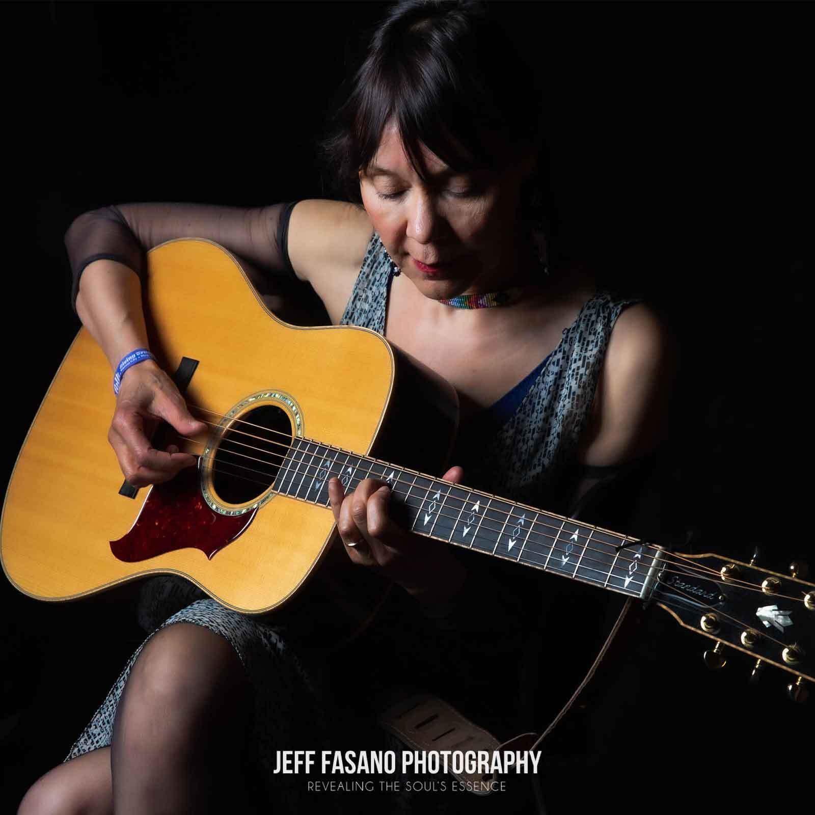 A woman sits and plays an acoustic guitar, looking down at the instrument, reflecting the spirit of Indigenous music. She is illuminated against a dark background. "JEFF FASANO PHOTOGRAPHY" text is displayed at the bottom of the image.