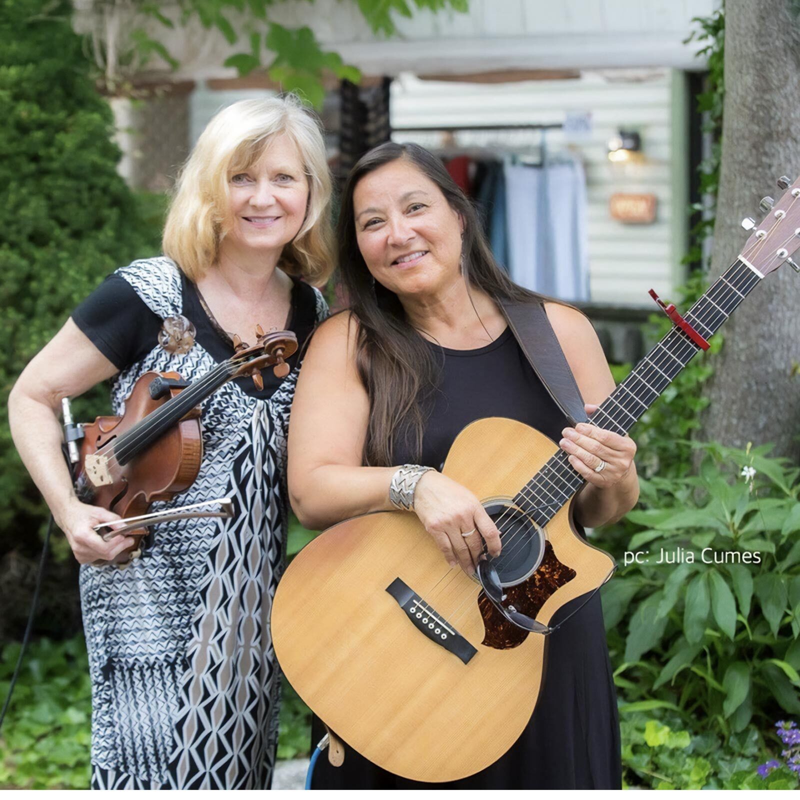 Two women standing outdoors, one holding a violin and bow, the other an acoustic guitar. Smiling amid greenery with a building behind them, they appear ready to share Indigenous music at a vibrant live music event.