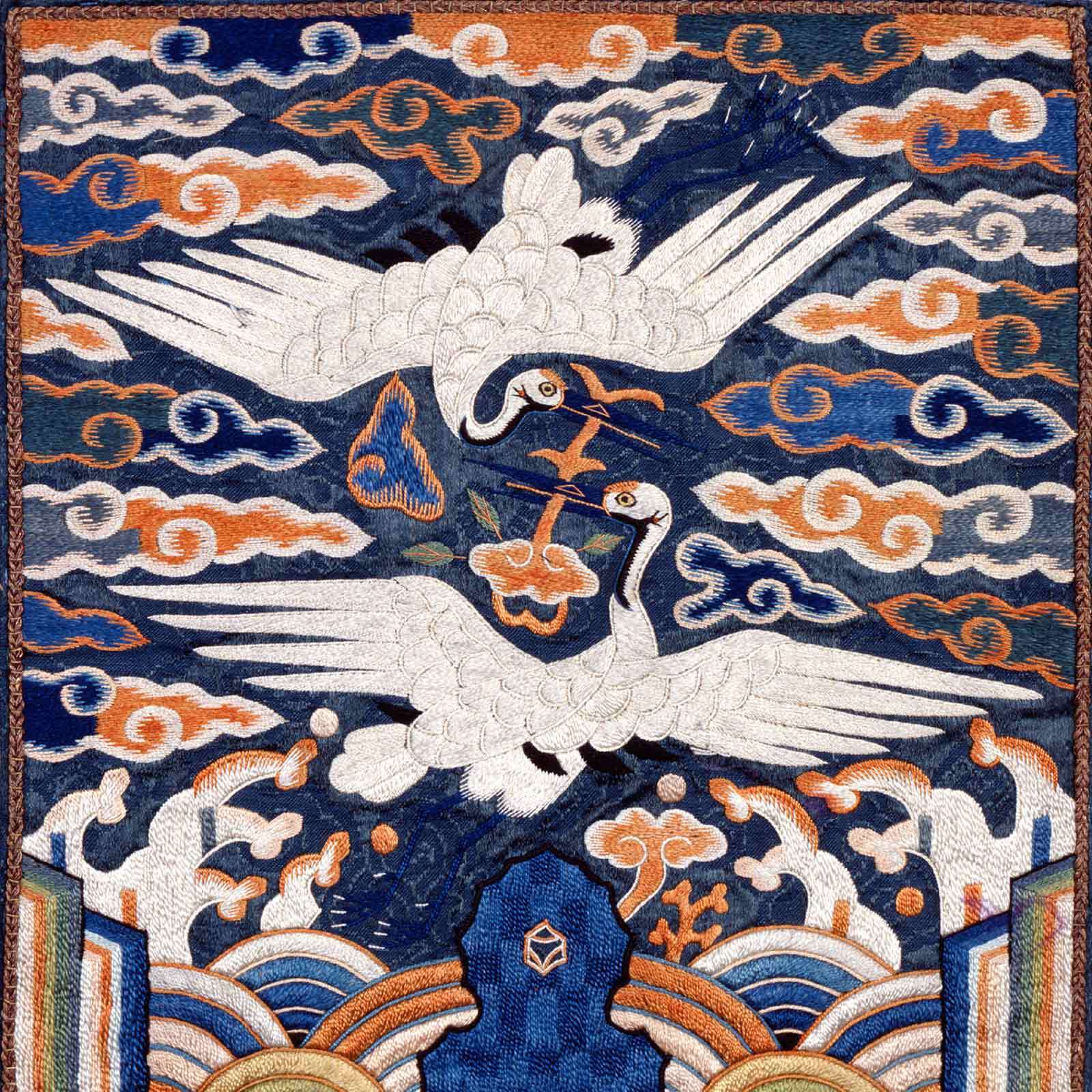 A detailed embroidered panel of Korean art shows two white cranes facing each other in flight above stylized clouds and waves, with blue, orange, and white colors dominating the scene. Perfect for a Members-Only Tour experience.