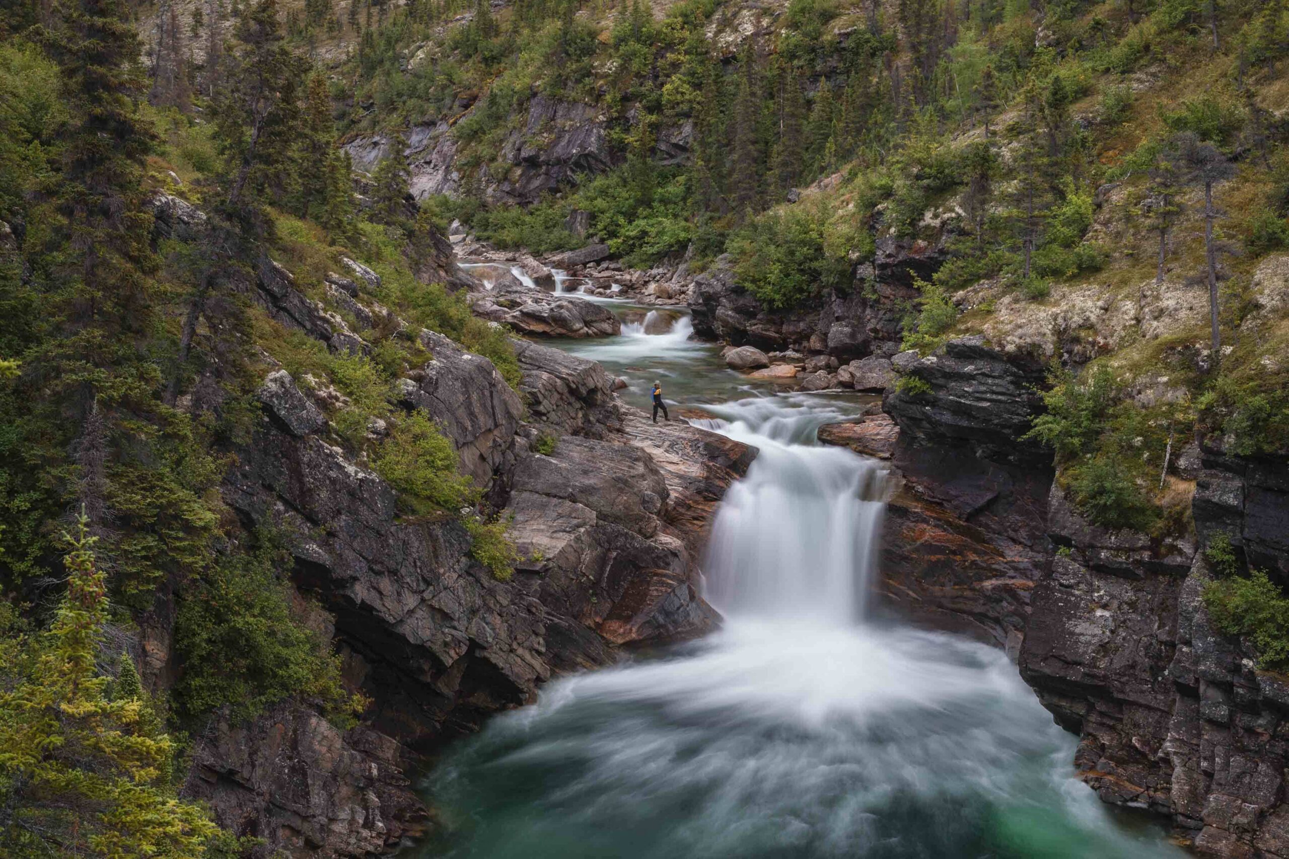 A person stands on a rocky ledge above a waterfall surrounded by forested cliffs and flowing water, with trees and greenery covering the area.