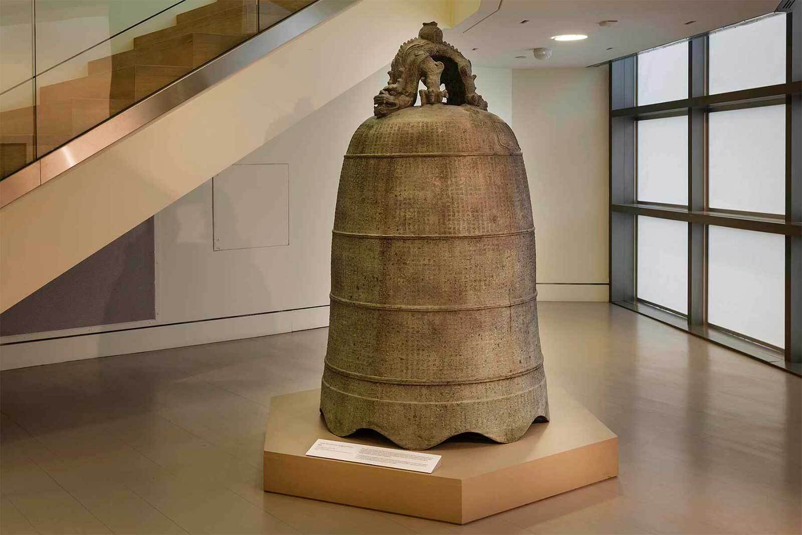 A large bronze bell with a decorative dragon-shaped handle, symbolizing Double Happiness in Chinese art, is displayed on a raised platform in a modern museum setting near a glass wall and staircase.