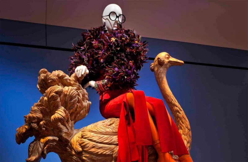 A mannequin wearing a black feathered top, red pants, and large round glasses is seated on an ornate wooden sculpture of a bird against a blue and beige background.