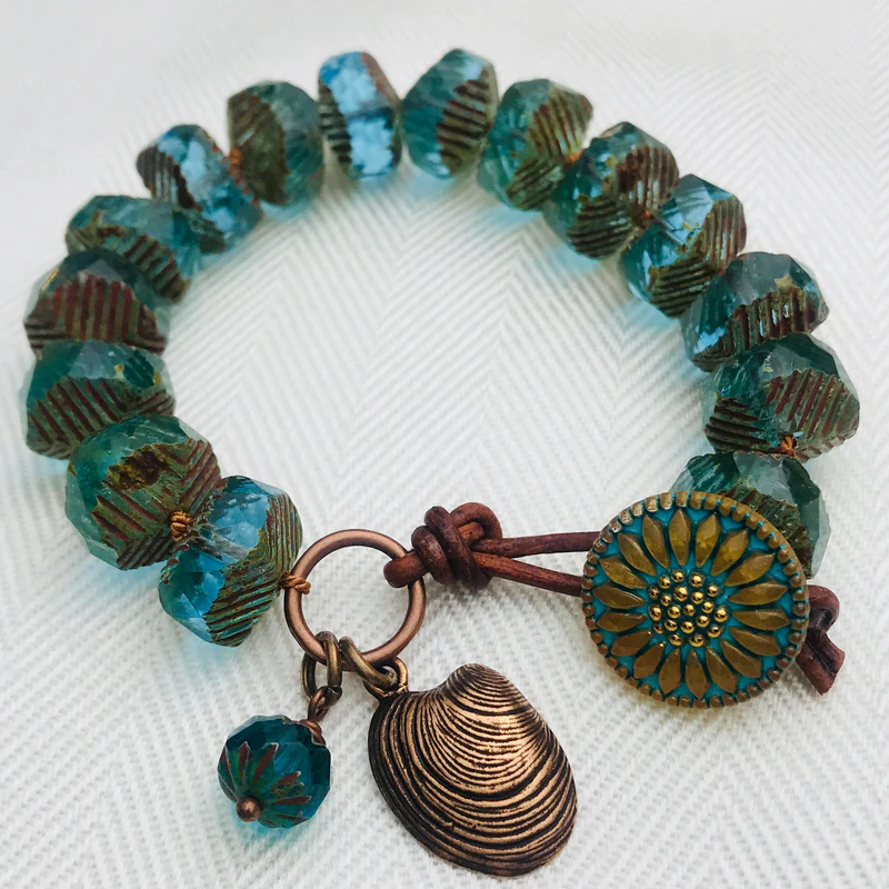 A bead wrap bracelet made of faceted turquoise glass beads with bronze accents, featuring a sunflower-patterned button clasp, a bronze shell charm, and a small matching bead charm, all displayed on a white textured background.