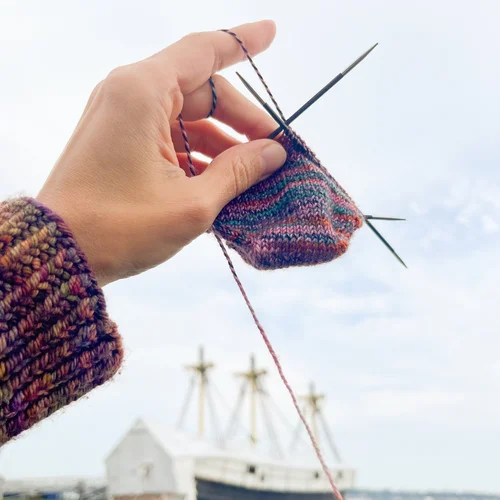 A hand in a multicolored knit sleeve holds knitting needles, stitching a small, vibrant project. In the background, ships with tall masts line the wharf beneath a cloudy sky.