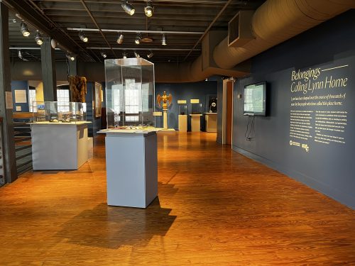 A museum exhibit room with wooden floors, blue display pedestals, and artifacts—belongings from the Calling Lynn Home Exhibit—are showcased in glass cases. Informational text lines the walls as track lighting illuminates each display.