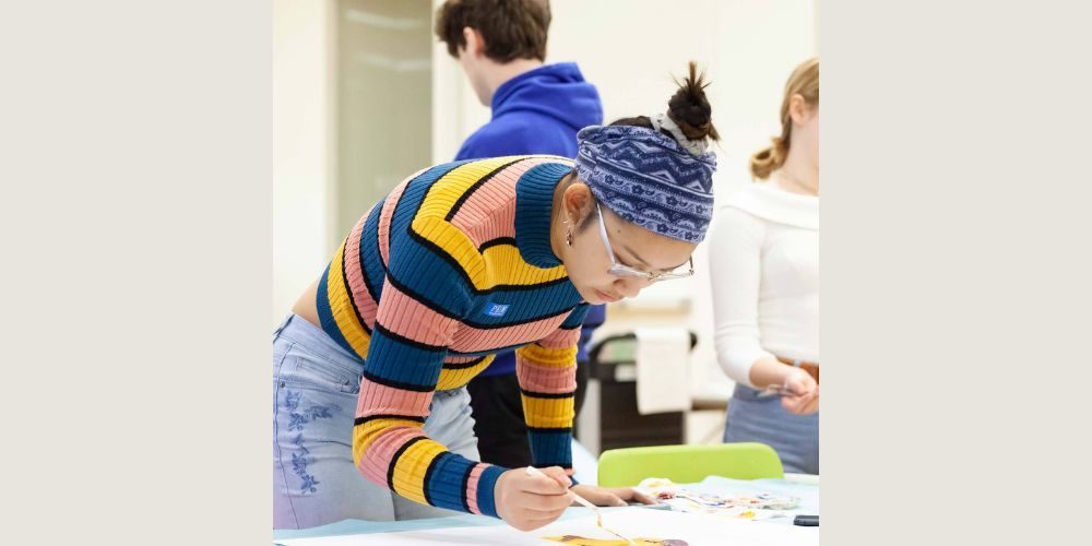 A person wearing a striped sweater and patterned headband leans over a table, focused on painting or drawing. Two others stand nearby, partially visible. The setting appears to be a classroom or studio.