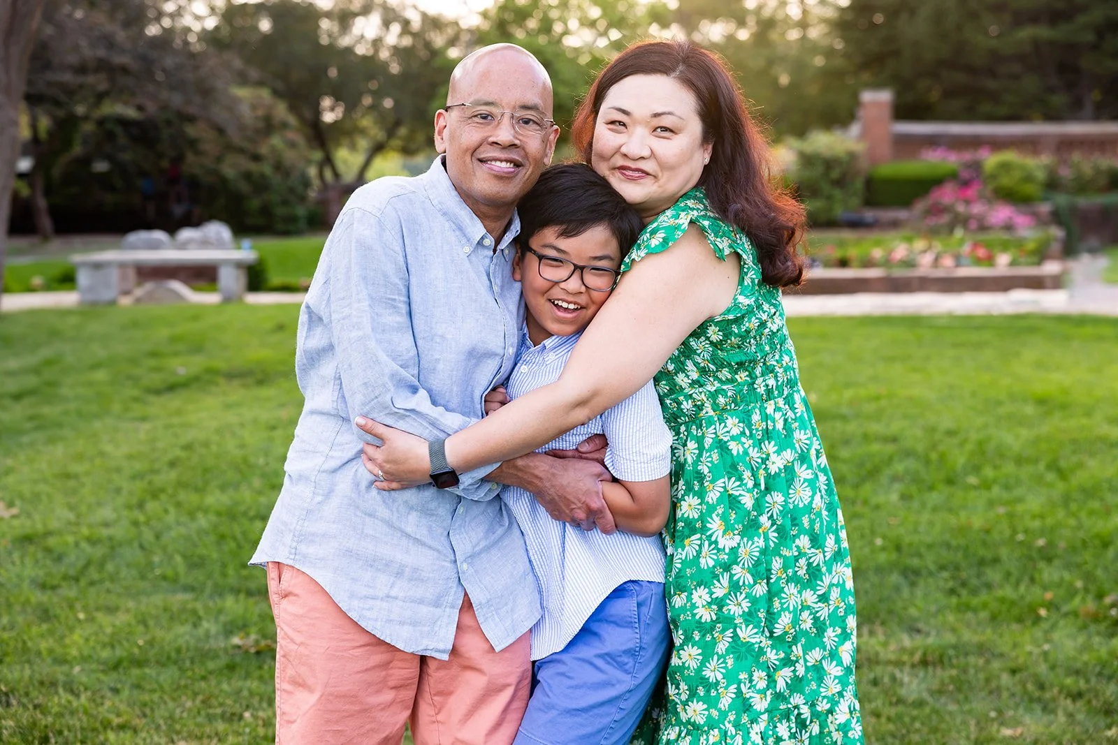 A man, woman, and young boy are standing outside on grass at Lynch Park, smiling and hugging each other. Perfect for family photography or mini sessions, the man wears a light blue shirt and pink shorts; the woman is in a green dress, and the boy in blue.