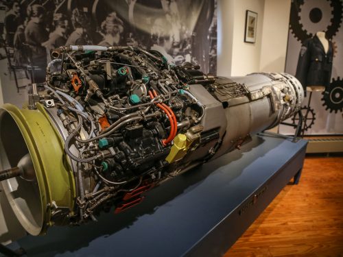 A detailed view of a jet engine on display in a museum setting, with visible wiring and components; historical photos, a suit, and industrial gear decorations are in the background.
