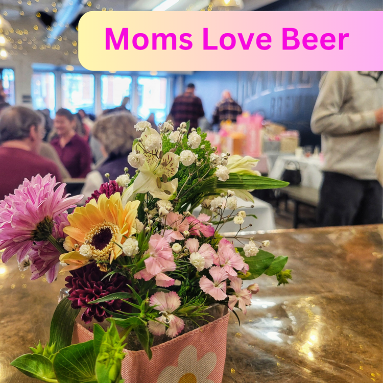 A close-up of a colorful flower arrangement on a table in a busy restaurant, with people seated at tables. Text at the top reads, "Moms Love Beer" in bold letters, celebrating moms and their passion for beer curated flights.